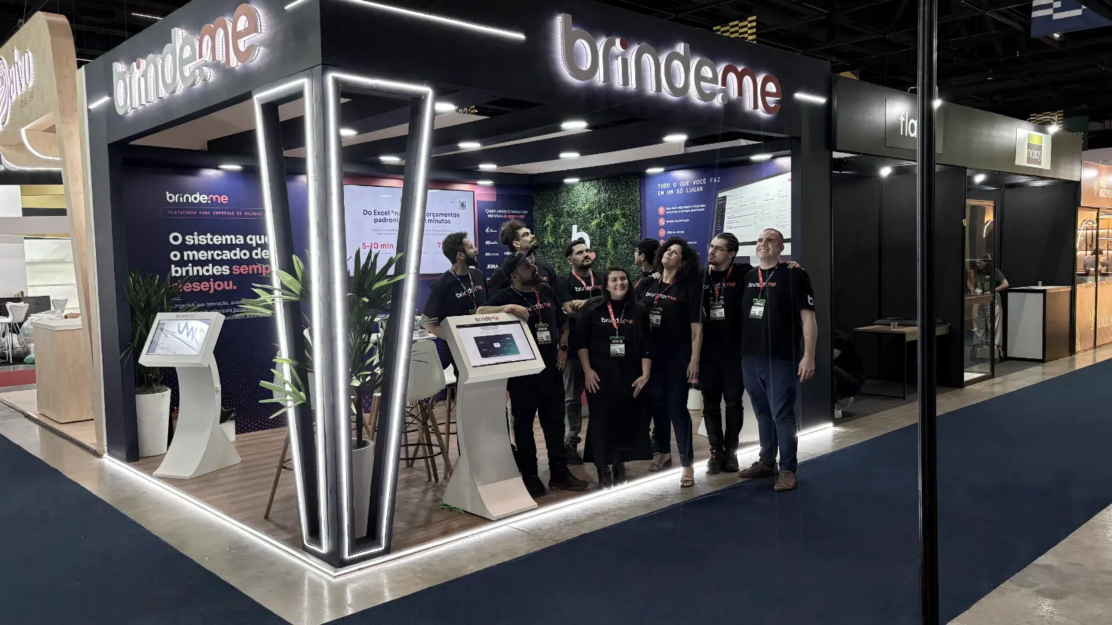 Estande da Brinde.me em feira do setor de brindes, com o time reunido e totens de demonstração